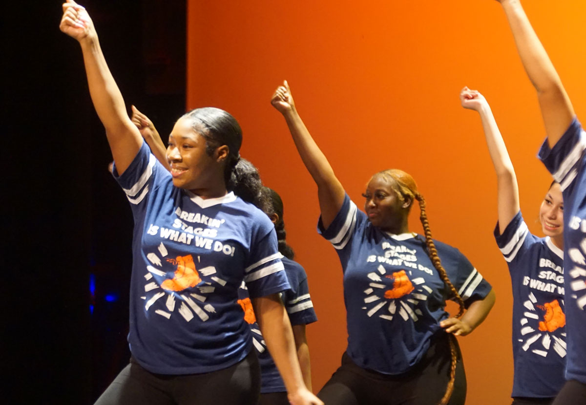 A Garland ISD STEP team performing a routine on stage, with the performers smiling while lifting their right hands in the air in a fist.