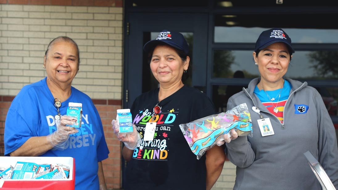 Three smiling lunch ladies posing with milk cartons and food outside a school.