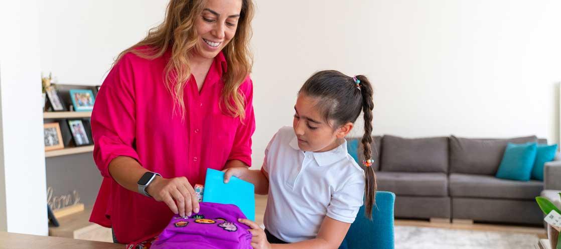 Mother helping young elementary age daughter put school supplies in backpack