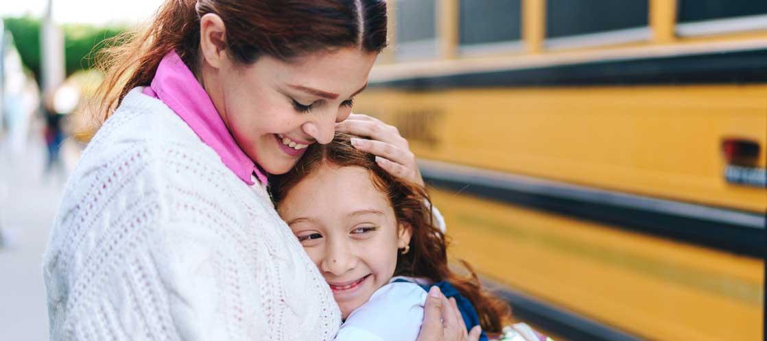 Mother hugging smiling young girl in front of school bus