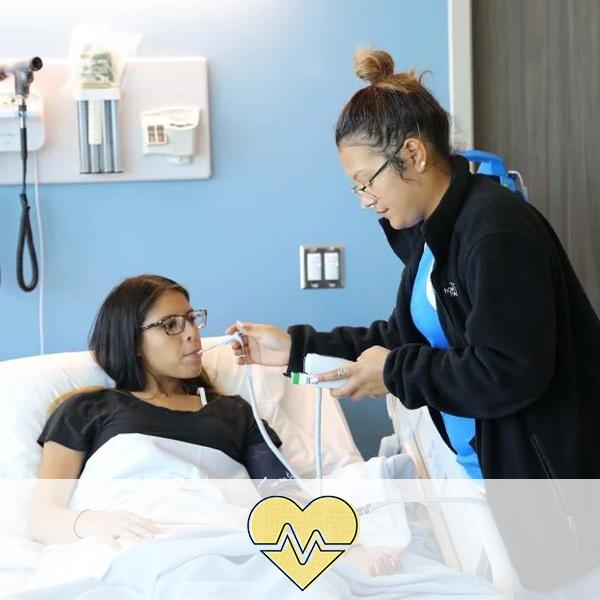 A student taking the blood pressure and temperature of another student in a hospital bed. A heart icon with a pulse rate is overlaid on the image, representing the Health Sciences cluster.