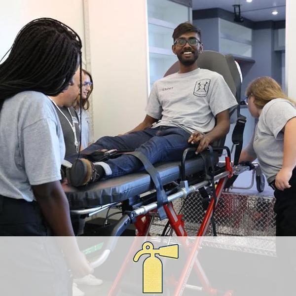 Students practicing loading people into an ambulance. A fire extinguisher icon representing the Law & Public Service cluster is overlaid on the image.