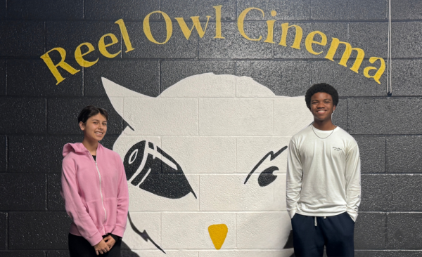 One female and one male student stand in front of a wall with the Reel Owl Cinema logo 