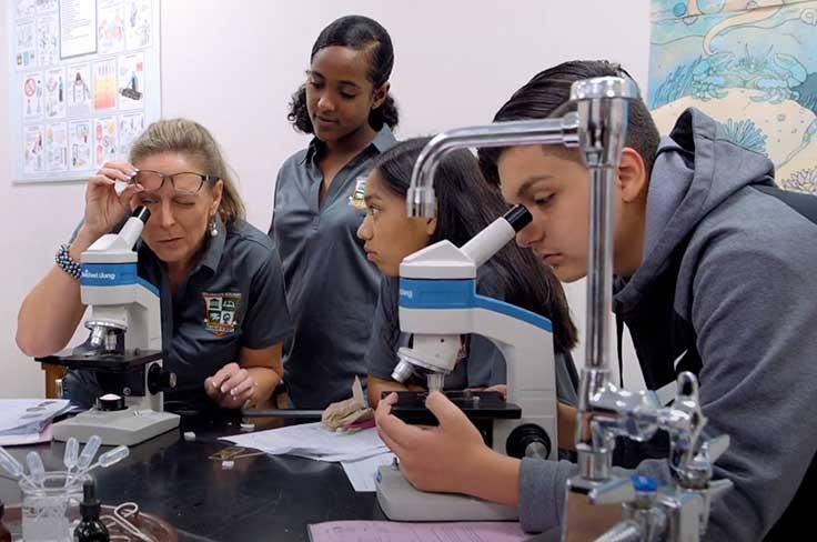 HS teacher and student looking through microscopes while other students look on.