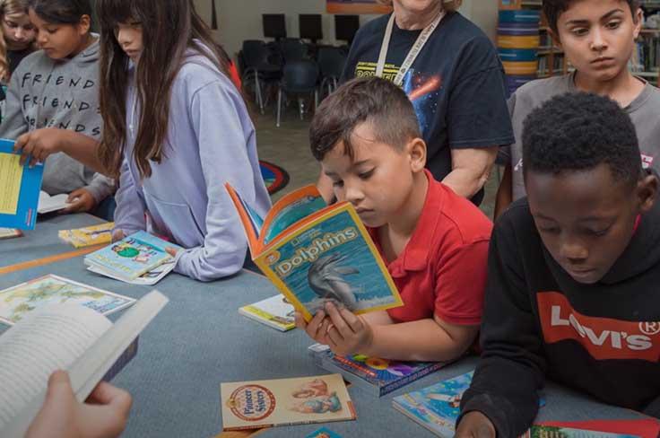 Young boy reading magazine surrounded by other students looking through books on a table.