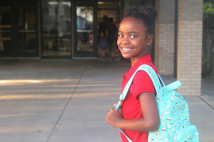 Girl with wearing backpack smiling as she pauses near school entrance.