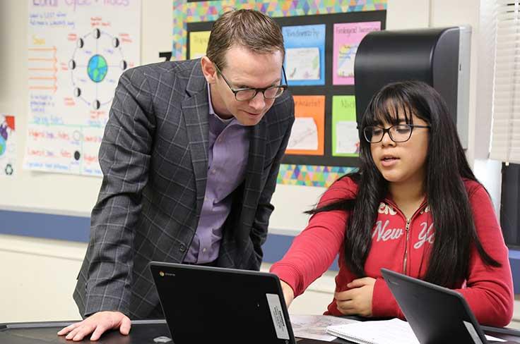 Girl student pointing at device screen while teacher leans over to assist.