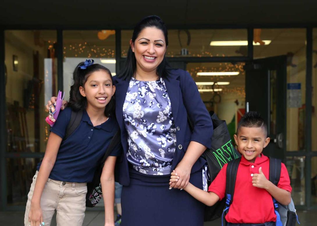 A smiling mother posing with her two excited students in front of a school building