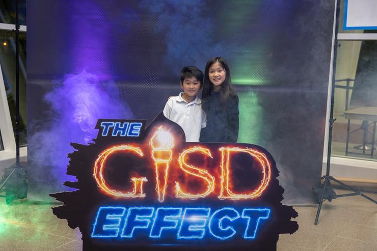 An Asian boy and girl hug and smile behind The GISD Effect sign