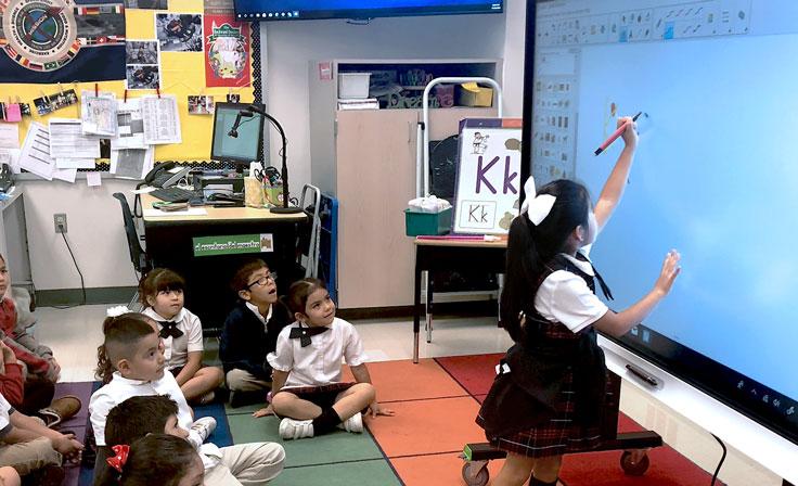 A young student writing on a smart board in class, with an overhead projector, computer and TV in the background.