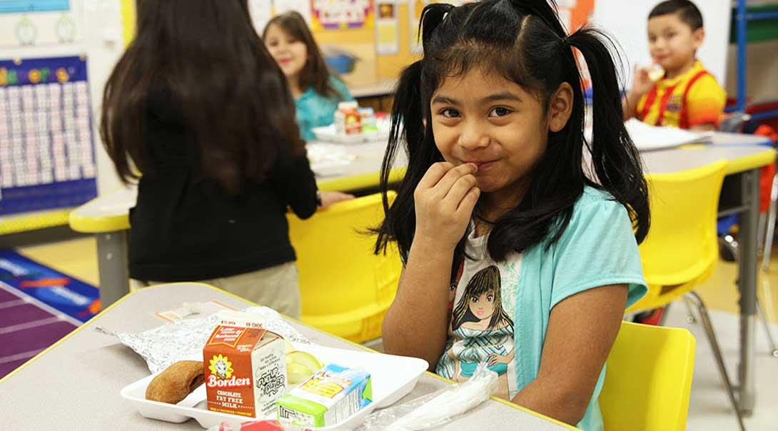 A girl smiling while eating breakfast at her desk.