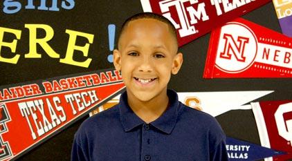 A young student smiling in front of a backdrop of college flags.
