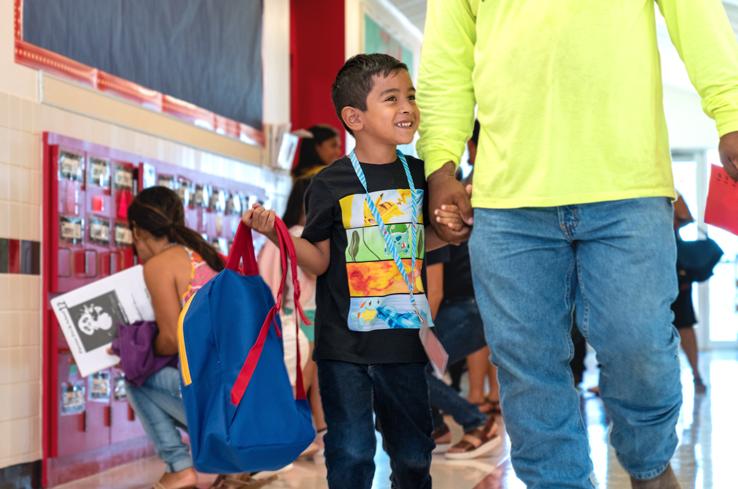 A prekindergarten student holding his father's hand and a backpack in the other while walking down a hall.