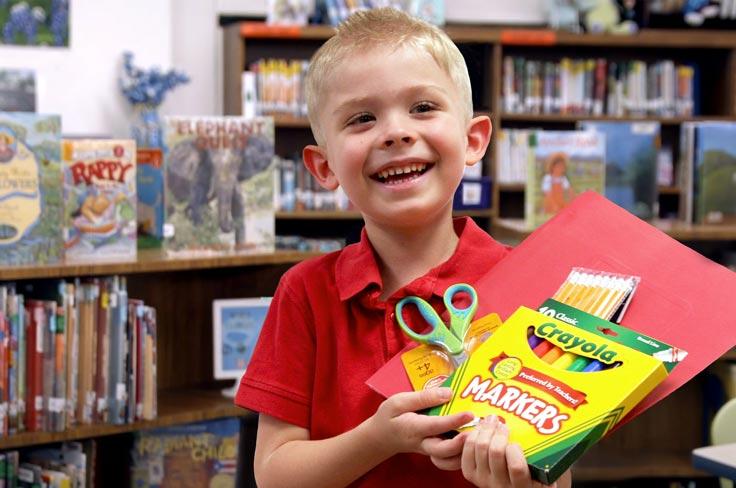 A student smiling in a library holding markers, scissors, a folder, and pencils for school.