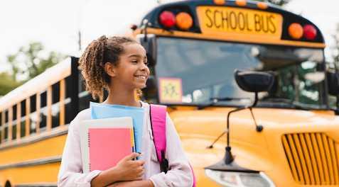 A girl smiling holding supplies while walking from a school bus.