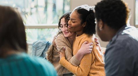 Two girls side hugging each other in a hallway, one in a yellow sweater.