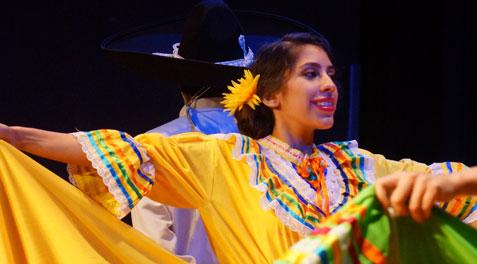 A closeup of a high school girl in bright and colorful green and yellow ruffled dress, smiling and dancing Folklorico with her team.