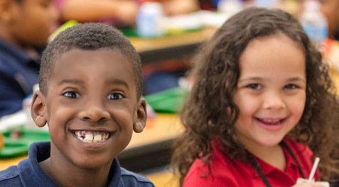 Two smiling kids at a lunch table in the cafeteria.