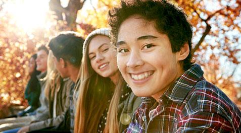 A row of teenagers sitting outside in long-sleeved shirts, with trees dropping orange leaves behind them.