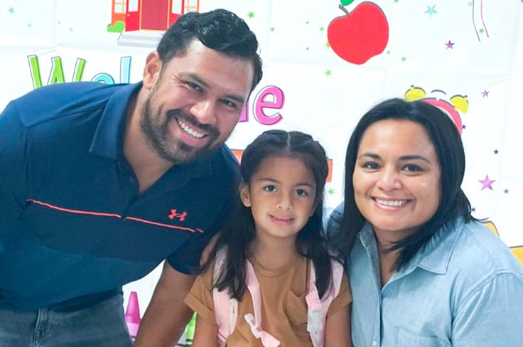 A mom and a dad smiling with their little girl in front of a "Welcome to Pre-K" backdrop.