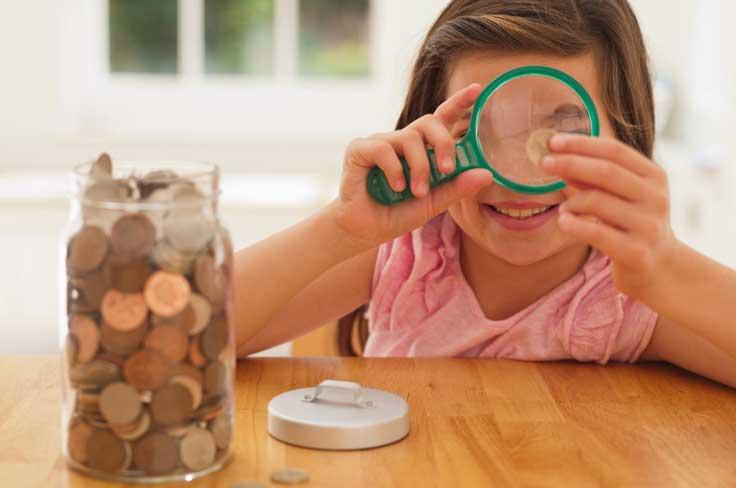 Girl looking through magnifying glass at a coin, with jar full of coins nearby