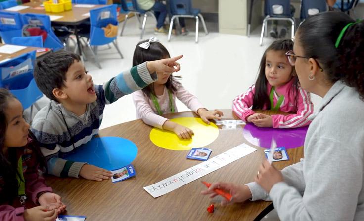 A prekindergarten teacher leading a small group with excited students. One of the students points to the board behind the teacher with a smile.