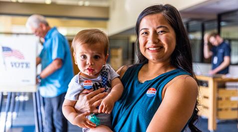 A Filipino woman holding her child while sporting an "I voted" sticker. In the background, an older Caucasian man casts his vote.
