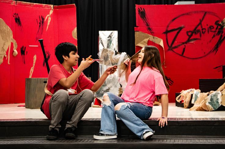 A middle school theatre production, featuring a red background with "no zombies" painted on it. A student dressed in gear wields scissors and grabs at another students hair to cut it, while the other student recoils.