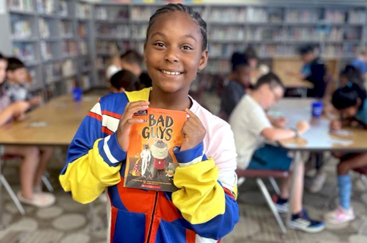 A young smiling student holding a book titled "The Bad Guys".