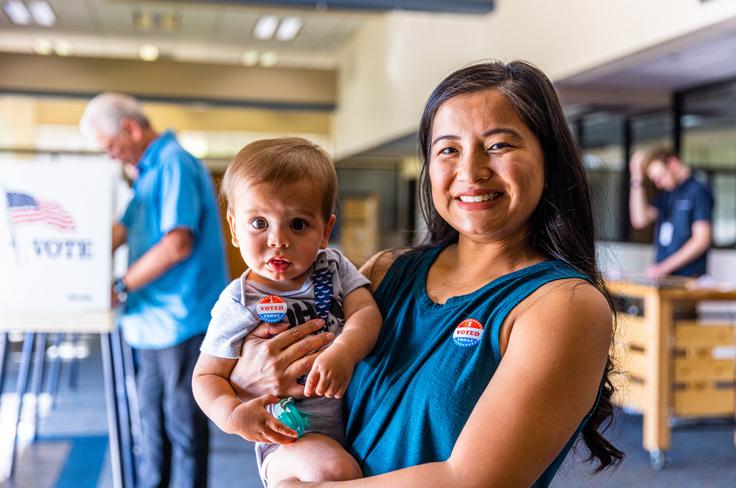 A Filipino woman holding her child while sporting an "I voted" sticker. In the background, an older Caucasian man casts his vote.