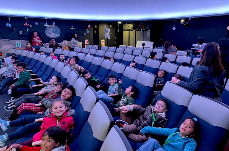 Young students grin with excitement and make silly faces while sitting in the planetarium under the projection.