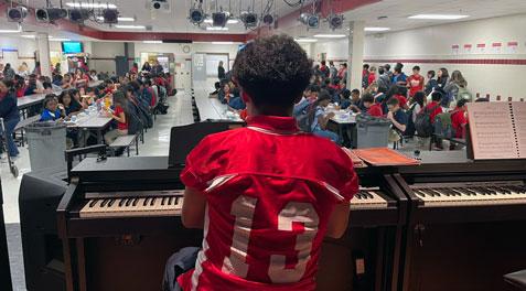 A student in a football jersey playing the piano for fellow students in the cafeteria.