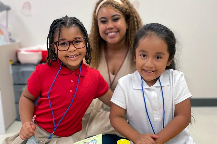 Two elementary girls posing together at a table with their teacher smiling behind them.