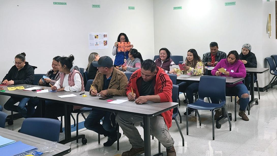 A class of adults works on a project using paper, pencil, colored paper and scissors.