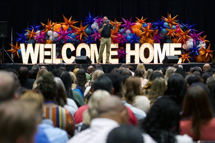 Dr. Adams addressing a crowd of new employees in front of a background of colorful balloons and large light-up letters stating "Welcome Home".