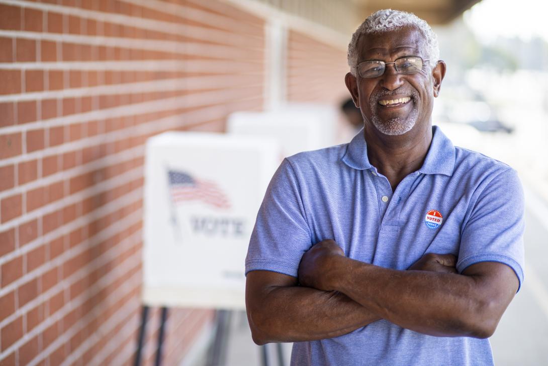 An African American gentleman smiling in front of voting booths with a "I Voted" sticker.