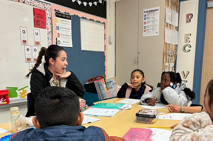 A teacher sits with elementary students at a round table while the students listen to her lesson attentively.