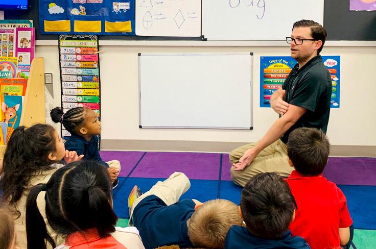 A prekindergarten teacher gestures to his arm as he sits on a carpet with his students.