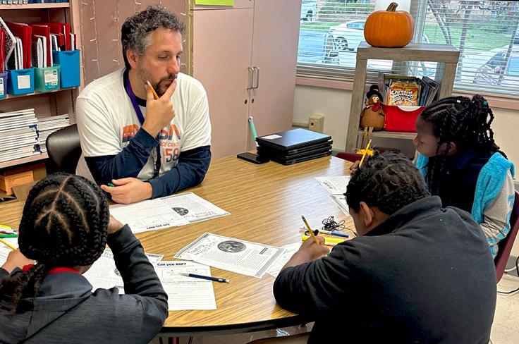 A teacher watches his students answer a question as they all work together on a group table assignment.