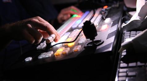 A closeup of a student using a soundboard at a district football game, the colors of the keyboard striking against the dark office.