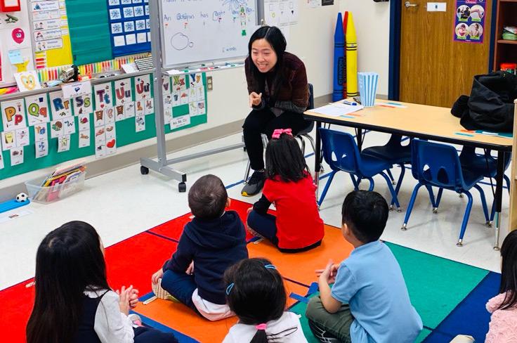 A teacher smiles and gestures, a group of students sitting attentively on the floor listening to her lesson.