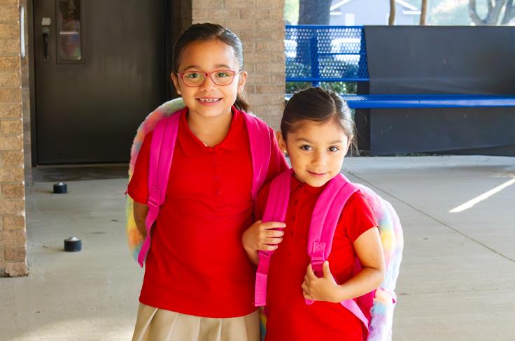 Two sisters posing together with their backpacks outside of an elementary school.