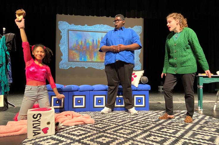 A smiling student raising her hand in pink and pigtails with two other students acting as surprised parents in the winning One Act Play, Sylvia.