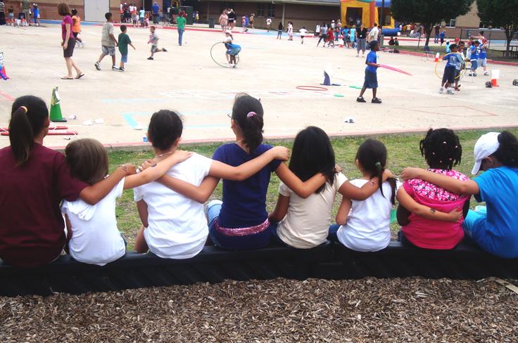 Group of kids sitting arm-in-arm watching other students play during recess.