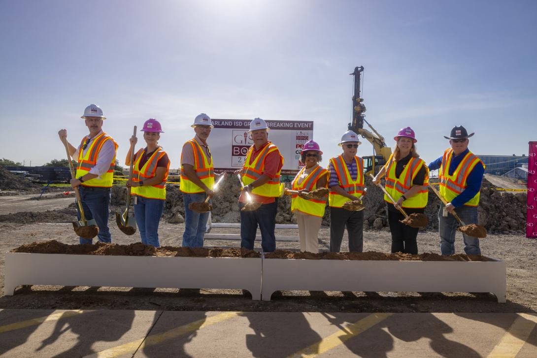 A group of adults stand in hard hats and vests with shovels extended
