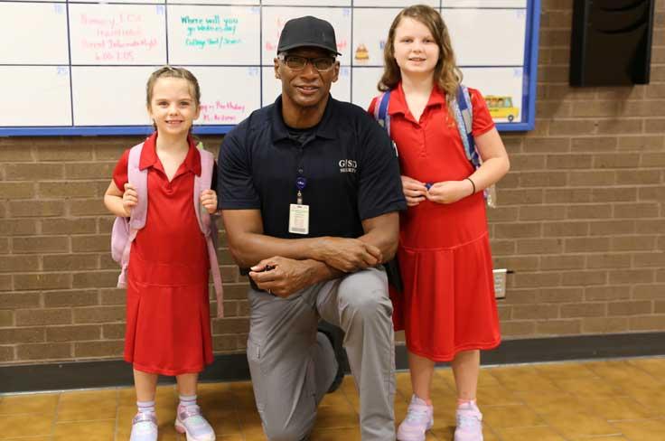 Armed security officer kneeling between two elementary girl students in front of school wall calendar