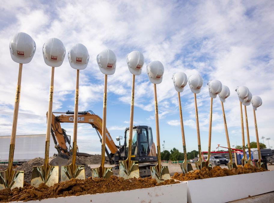 With construction equipment in the background, a row of helmets on shovel handles stand ready for the RHS MAC groundbreaking ceremony.