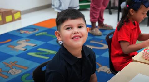 A young student smiles in a chair at a desk.
