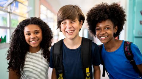 Three secondary students smiling in a school hallway with their backpacks.