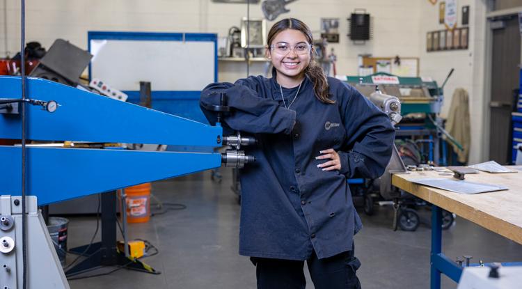 A girl wearing safety glasses and a uniform poses next to a manufacturing machine, smiling.